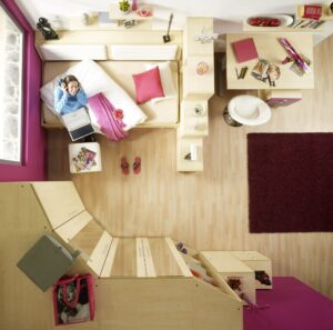 Jugendzimmer mit Holzmöbeln und pinken Wänden, Mädchen lächelt und schaut dabei in die Kamera