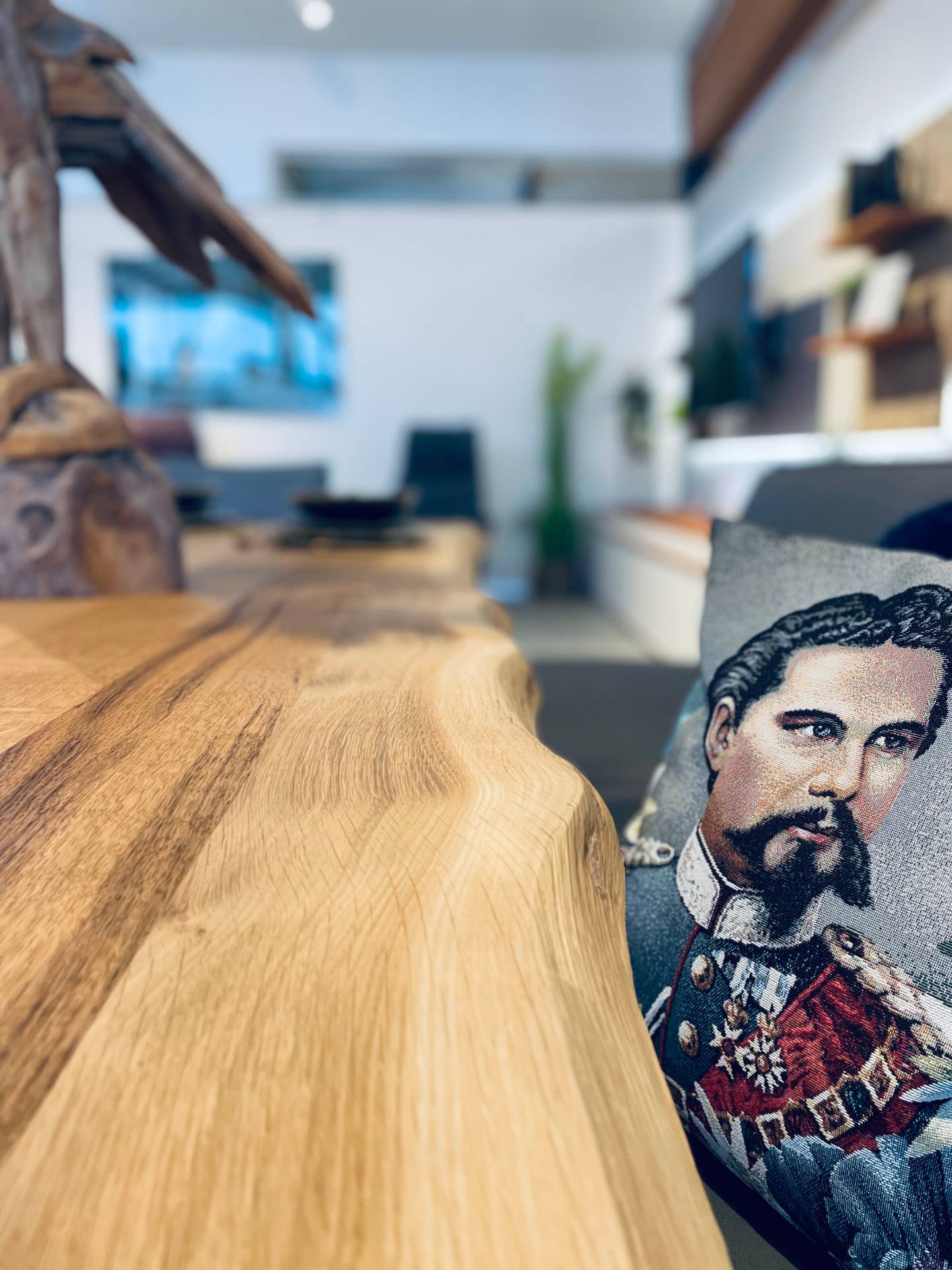 Nahaufnahme einer Massivholz-Esstischkante mit Baumkante und ein Kissen mit Porträt-Stickerei im Hintergrund.