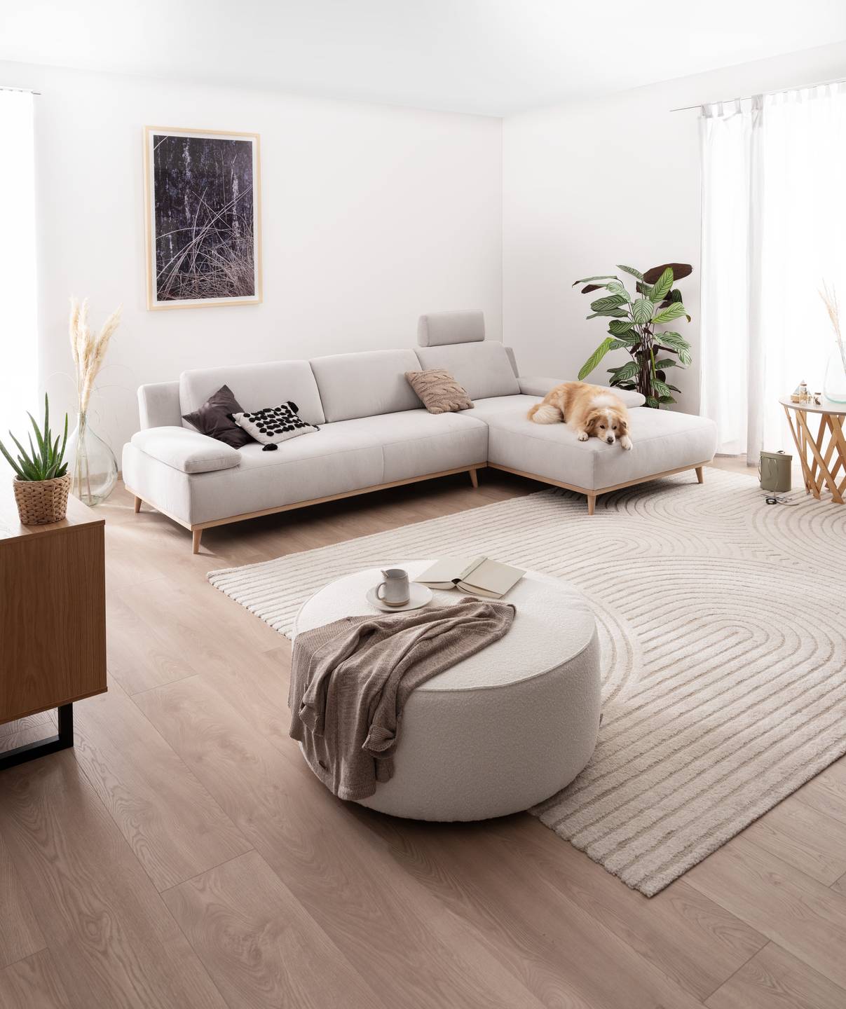 Modernes Wohnzimmer mit hellem Polstermöbel in L-Form, rundem Hocker, Teppich und Hund – Wohntrends 2026 bei Möbel Pölt.